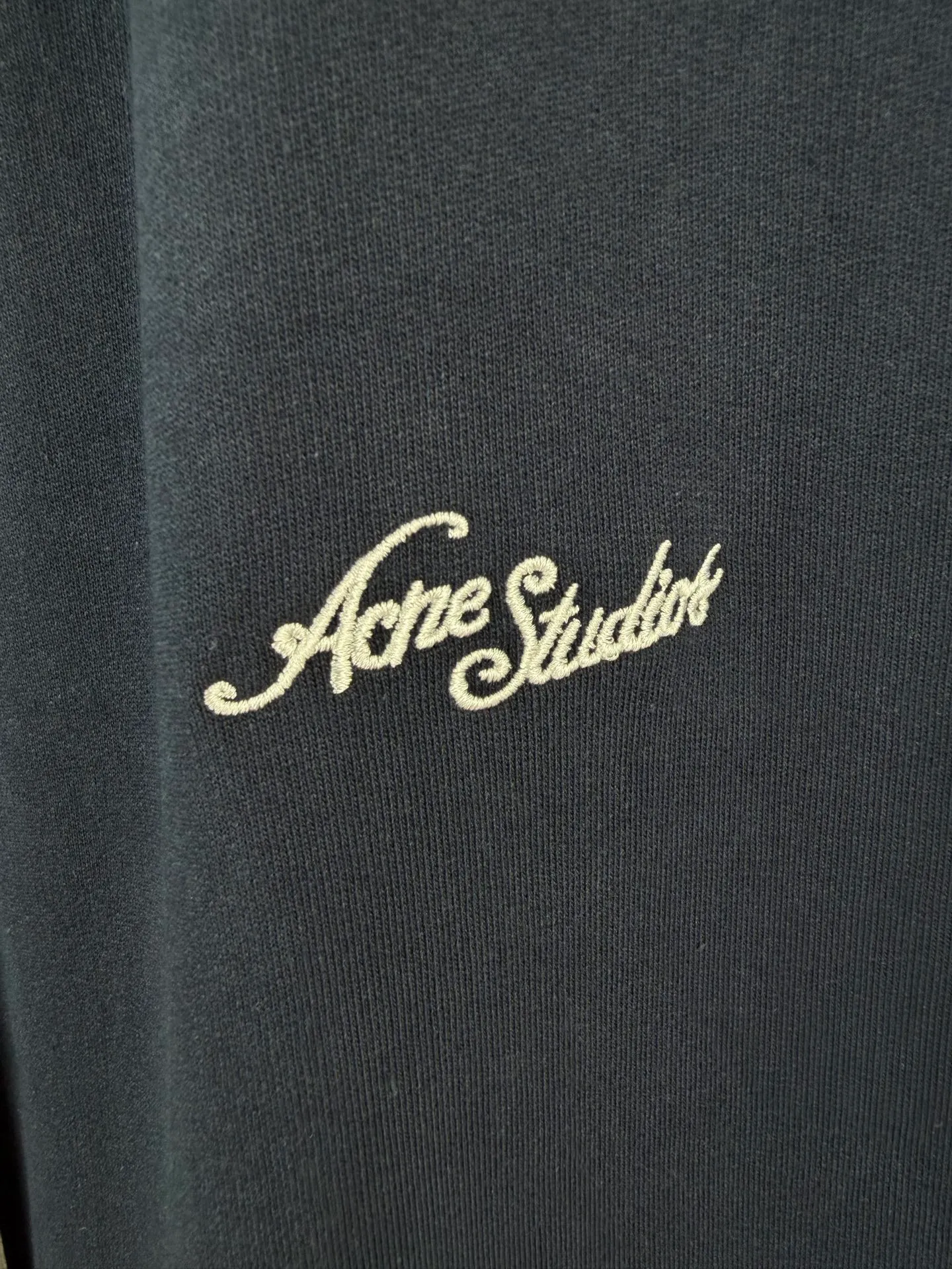 ACNE STUDIOS embroidered logo fleece sweatpants wide-leg trousers. Fabric: 32-count double yarn top + 16-count terry bottom yarn G The veil is made of 32-count fine yarn. The high-count yarn is smooth and delicate. After weaving, it forms a smooth and compact surface. It not only resists pilling and wear-resistant, but also gives the fabric a touch as delicate as skin. The base yarn is made of 16-count roving. The low-count yarn is fluffy and hairy. It naturally forms a rich terry layer during the weaving process, providing a sufficient source of fiber for subsequent sanding. This combination of fine surfaces and thick soles strikes a perfect balance between warmth and skin-friendliness. Mechanical sanding is the soul process of this fabric. By rubbing the bottom surface of the roller wrapped with emery leather at high speed, the fiber ends of the 16-count roving are evenly pulled out and cut off, forming a dense and uniform short pile layer. The smoothness and neatness of needle embroidery and the soft velvet feel of brushed fabrics form a material dialogue between hardness and softness. The embroidery area is smooth and crisp, with sharp lines, like delicate inscriptions carved on a rich suede canvas; while the surrounding brushed fabric presents a soft matte natural texture. The spacing between stitches is consistent and the strength is balanced to ensure that each stitch is placed accurately. This rigorous stitching structure makes the edges of the logo present a sharp and clear outline, and the strokes of the text are full and smooth. Even the curved turning points of the floral fonts can achieve a smooth transition through the connection of tight stitches.